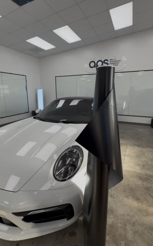 Roll of XPEL Satin Midnight Black color PPF next to a white Porsche 911